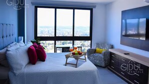 Un dormitorio moderno con un ventanal grande, una cama, un televisor y una mesa de comedor con fruta y una bebida.