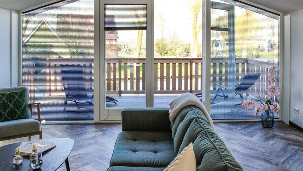 A cozy living room with a dark green sofa, a wooden coffee table, and a view through large glass doors to an outdoor deck with a wooden fence and chairs.