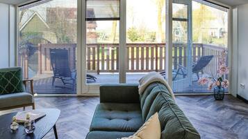 Een gezellige woonkamer met een donkergroene bank, een houten salontafel en een uitzicht door grote glazen deuren op een terras met houten omheining en stoelen.