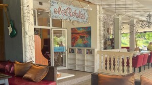 Un restaurante con un cartel que dice "Mo' Room Now", mesas al aire libre con cojines rojos y un cuadro colorido sobre la entrada.