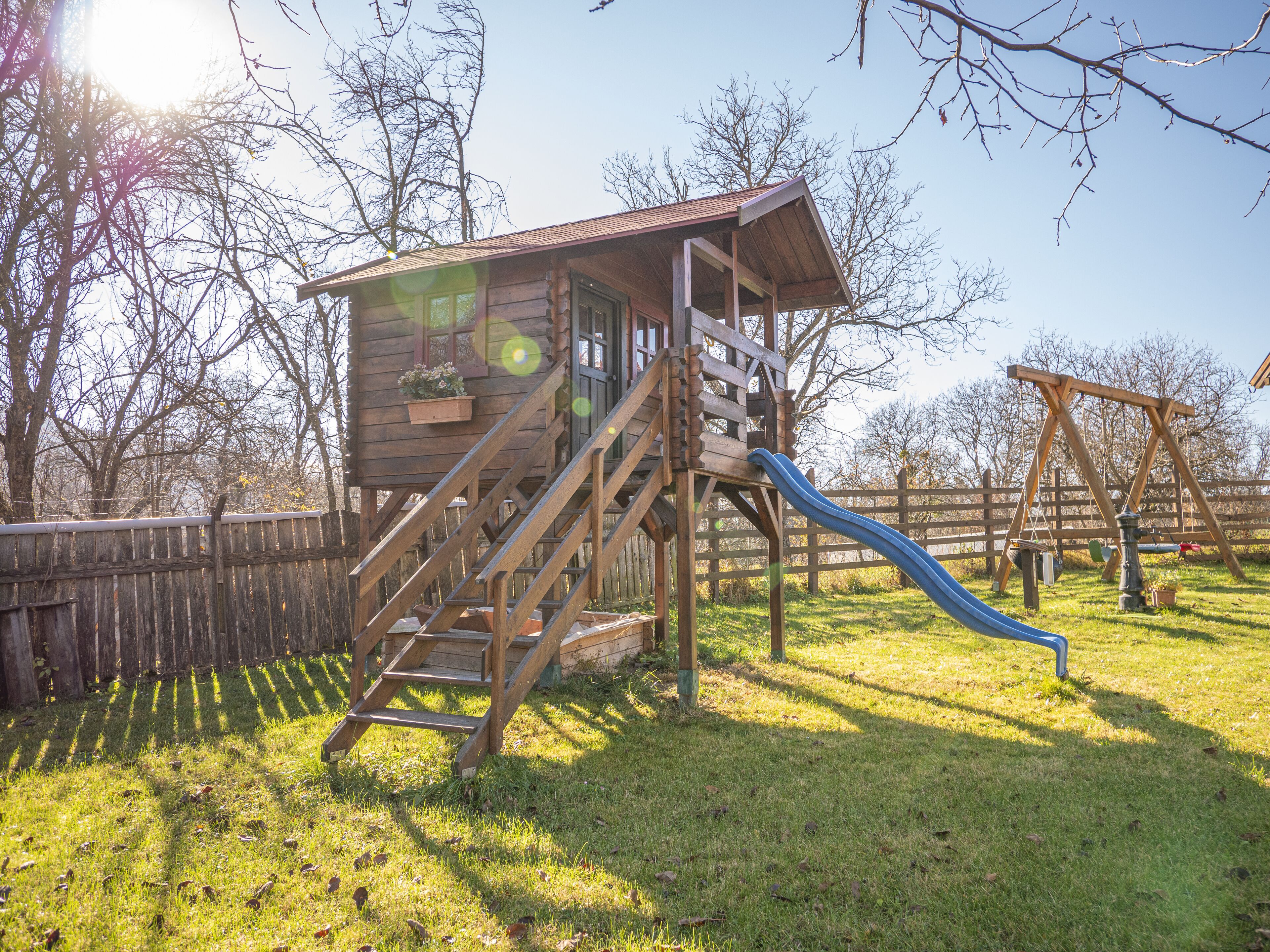 Une cabane en bois avec un toboggan, entourée d’une clôture en bois et d’arbres.