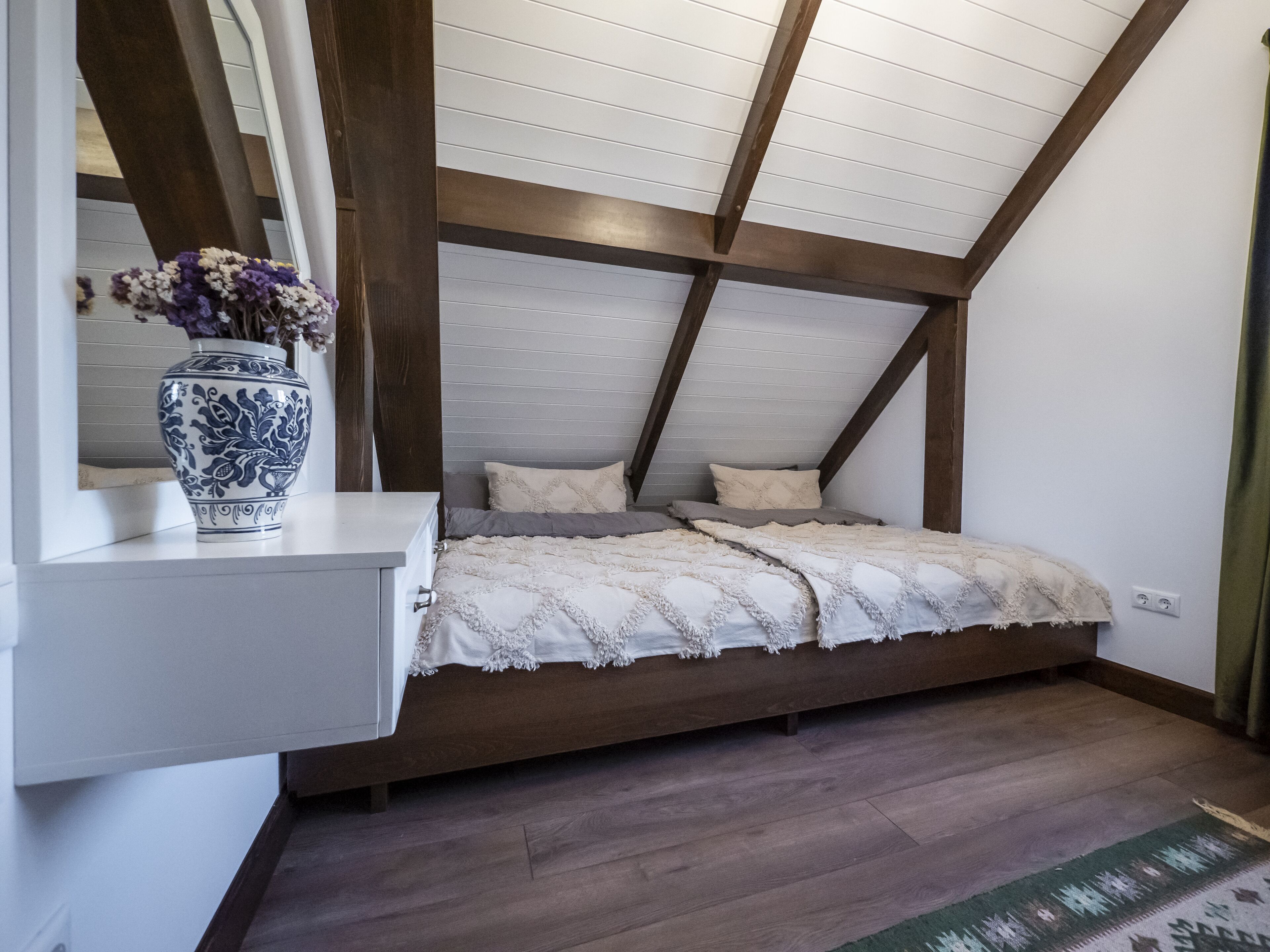 Ein Schlafzimmer mit schrägen Wänden, einem Bett mit weißer und grauer Bettdecke sowie einer Vase mit Blumen auf einer weißen Kommode.