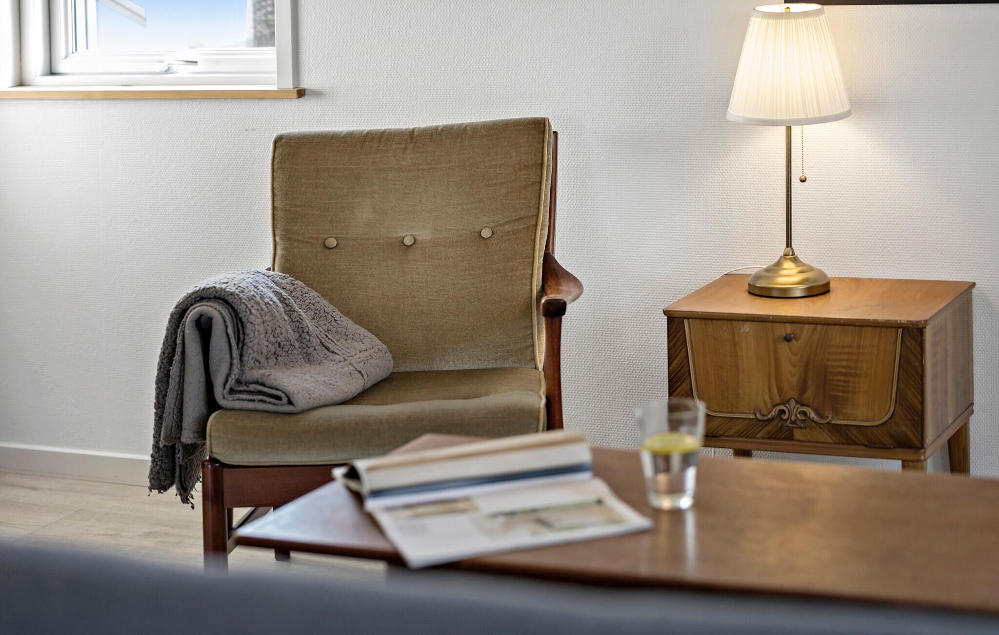 Une pièce confortable avec un fauteuil, une petite table de chevet en bois, une lampe et une table sur laquelle se trouvent des livres et un verre.