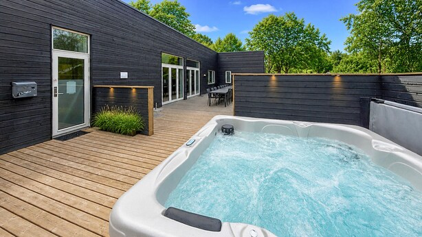 A modern outdoor hot tub area with a wooden deck, a glass door entrance, and a seating area with a table and chairs.