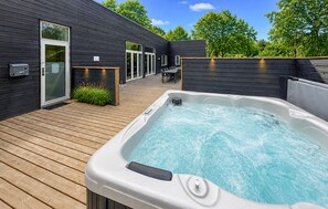A modern outdoor hot tub area with a wooden deck, a glass door entrance, and a seating area with a table and chairs.