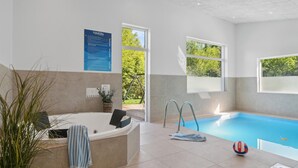 A modern indoor swimming pool with a jacuzzi, clear glass walls, and a view of greenery outside.