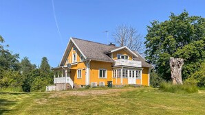Una casa amarilla de dos pisos con un balcón, rodeada de vegetación y un árbol grande.