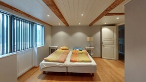 Un dormitorio con piso de madera, una cama con ropa de cama a rayas, una mesita de noche con lámpara y una ventana con persianas.