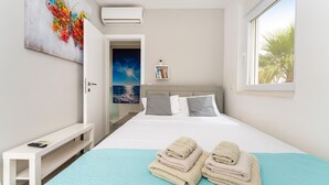 A bedroom with a bed, a bedside table, a wall-mounted air conditioner, a window with a view of palm trees, and a painting on the wall. - Stunning home in Zaton (Zaton)