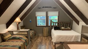 A cozy attic bedroom with a sloped ceiling, wooden beams, a bed with a white bedspread, two benches with patterned cushions, a small table with a lamp, and a window with a view of greenery. - Pine Ridge retreat on Beech Mountain, quiet with fireplace and mountain views! (Beech Mountain)