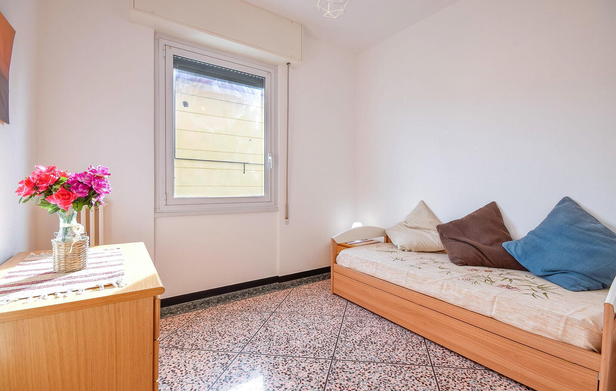 Quarto com cama, janela, escrivaninha com um vaso de flores e piso de azulejos.