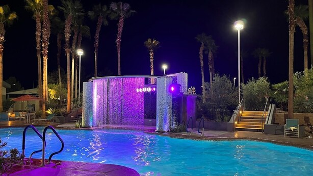 Zona de piscina nocturna con fuente de agua y palmeras.