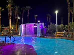 Un espace piscine nocturne agrémenté d’un élément décoratif aquatique et de palmiers.