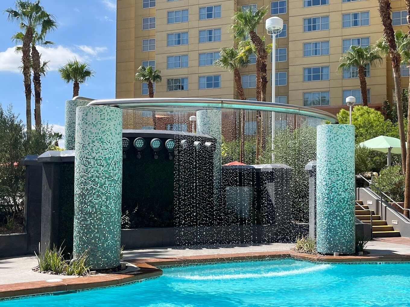 A swimming pool with a water feature, surrounded by palm trees and a building in the background.