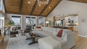 A spacious living room with a large sectional sofa, a coffee table, and a chandelier. There is a kitchen area visible in the background. - Mountain view’s with hot tub & pool table on Beech Mountain. Sleeps 10.  (Banner Elk)