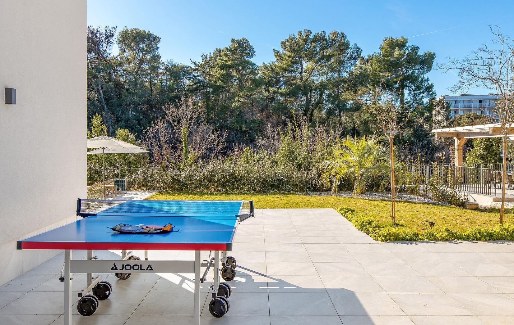 A ping pong table with a net, positioned on a tiled patio with a view of a forested area and a building in the background.
