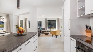 Una cocina moderna con armarios blancos, encimera oscura y un frutero. Hay una zona de comedor con un sofá y una mesa de centro.