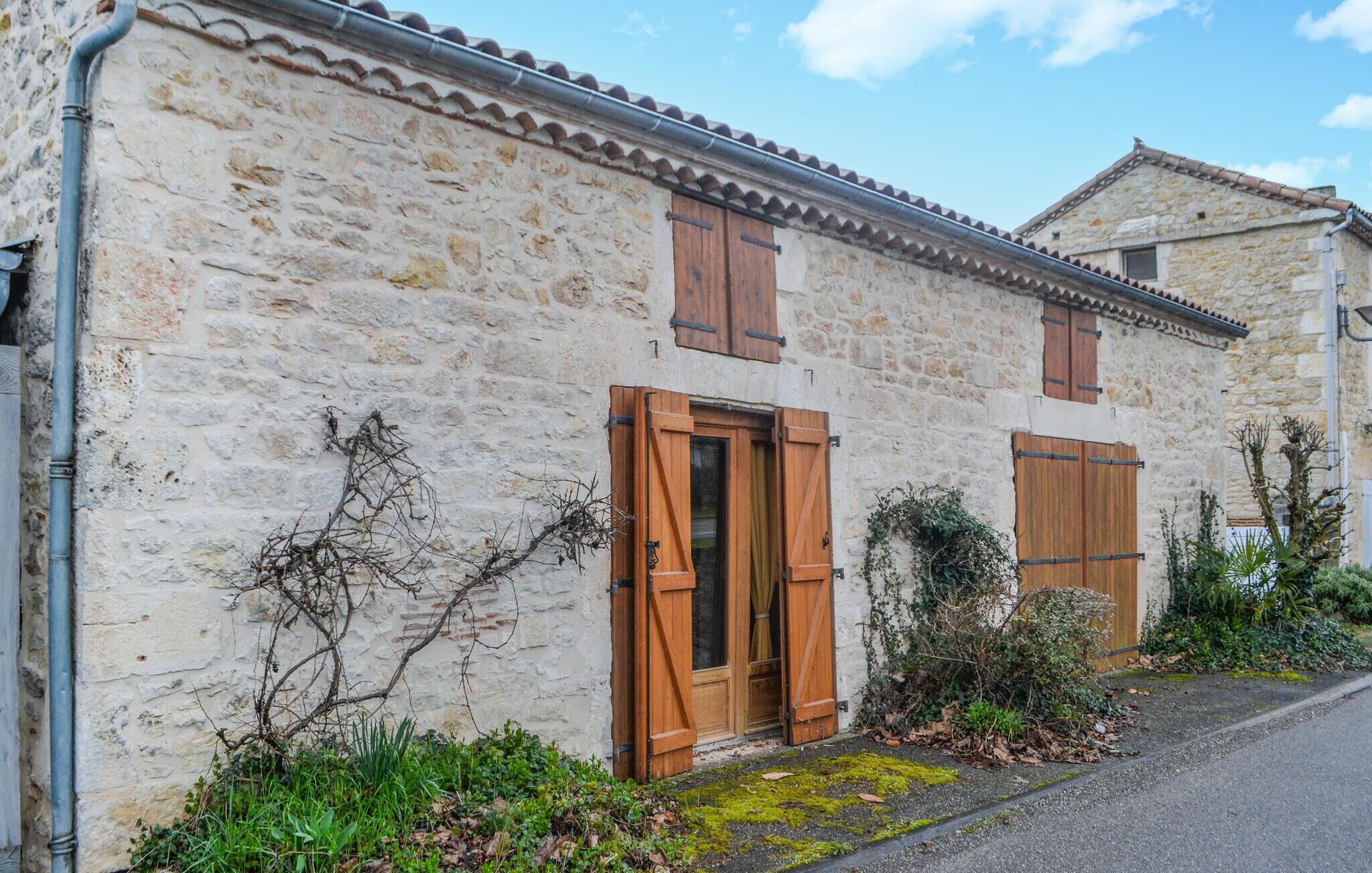 Un edificio de piedra con contraventanas de madera, una puerta visible y un pequeño jardín con plantas y un sendero.