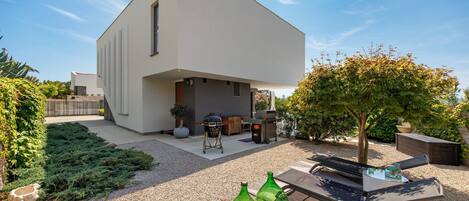 Una casa moderna con techo plano, un patio con parrilla y un jardín con un árbol y botellas decorativas.