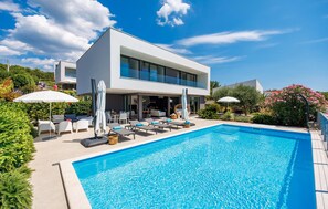 Une maison moderne à deux étages, dotée d’une piscine, d’un espace extérieur aménagé et d’une végétation luxuriante.