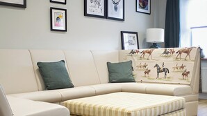 Un salon moderne avec un canapé beige, deux coussins verts, un pouf à rayures et un mur orné de tableaux encadrés.