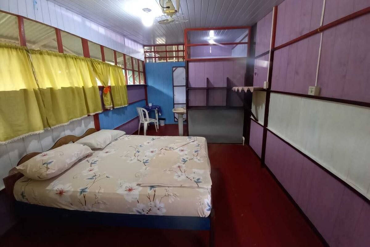 A room with a bed, a small table, and a chair. There is a window with curtains, and the walls are painted in different colors.