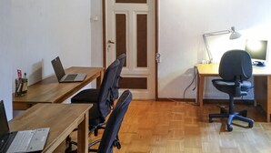 Una habitación con dos escritorios de madera, uno con un ordenador portátil y otro con un ordenador y monitor. Hay dos sillas de oficina negras, una lámpara y una puerta con marco marrón.