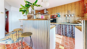 Une cuisine avec un mur en bois, un comptoir en métal ondulé, deux tabourets en osier et une plante en pot.