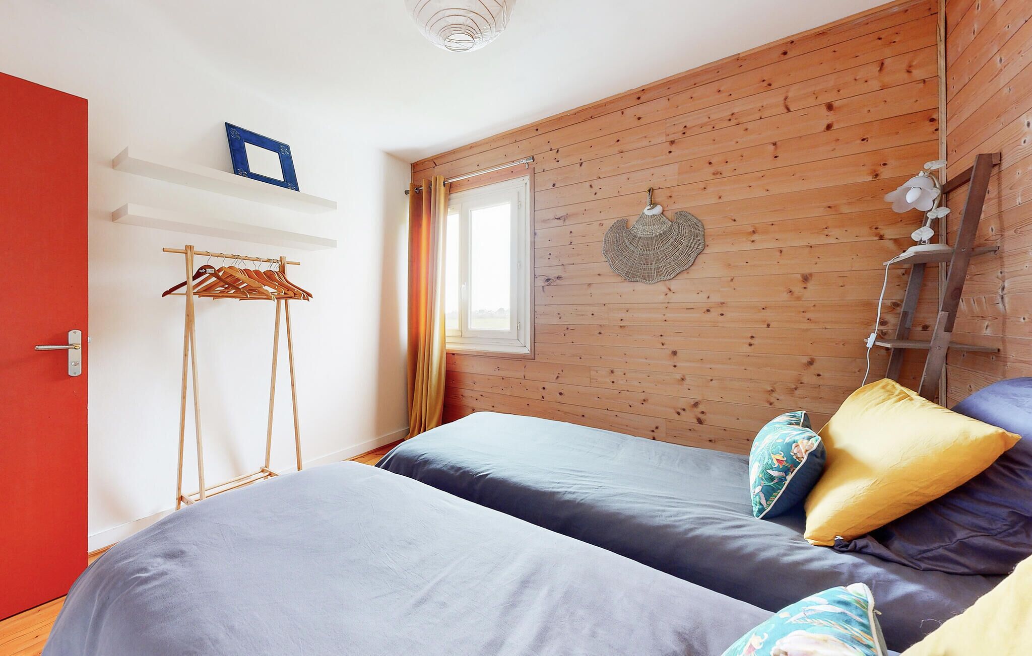A bedroom with two beds, wooden walls, a window, a hanging light fixture, and a door with a red frame.
