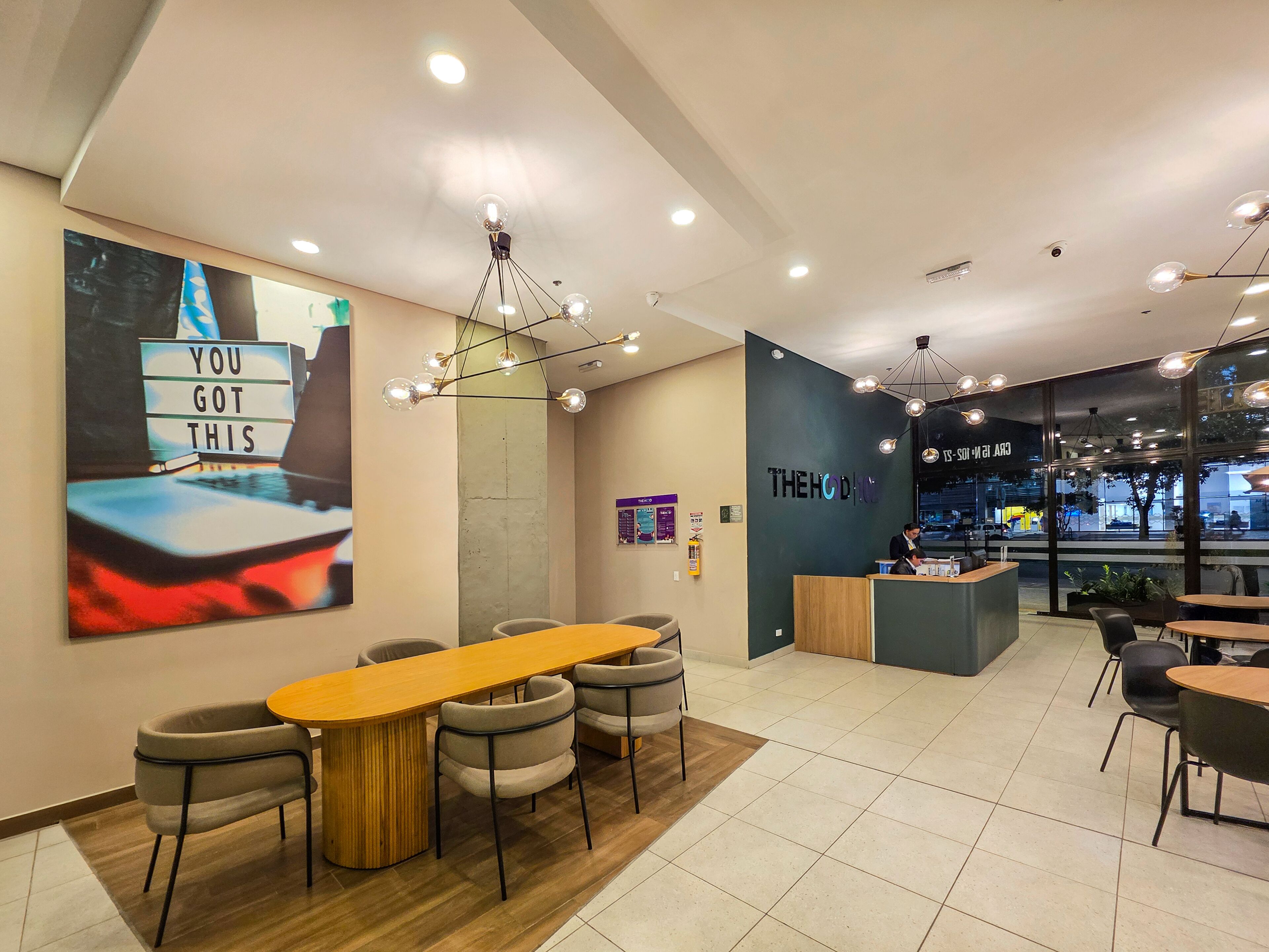 A modern cafe interior with a long wooden table, cushioned chairs, and a wall-mounted sign that reads "YOU GOT THIS".
