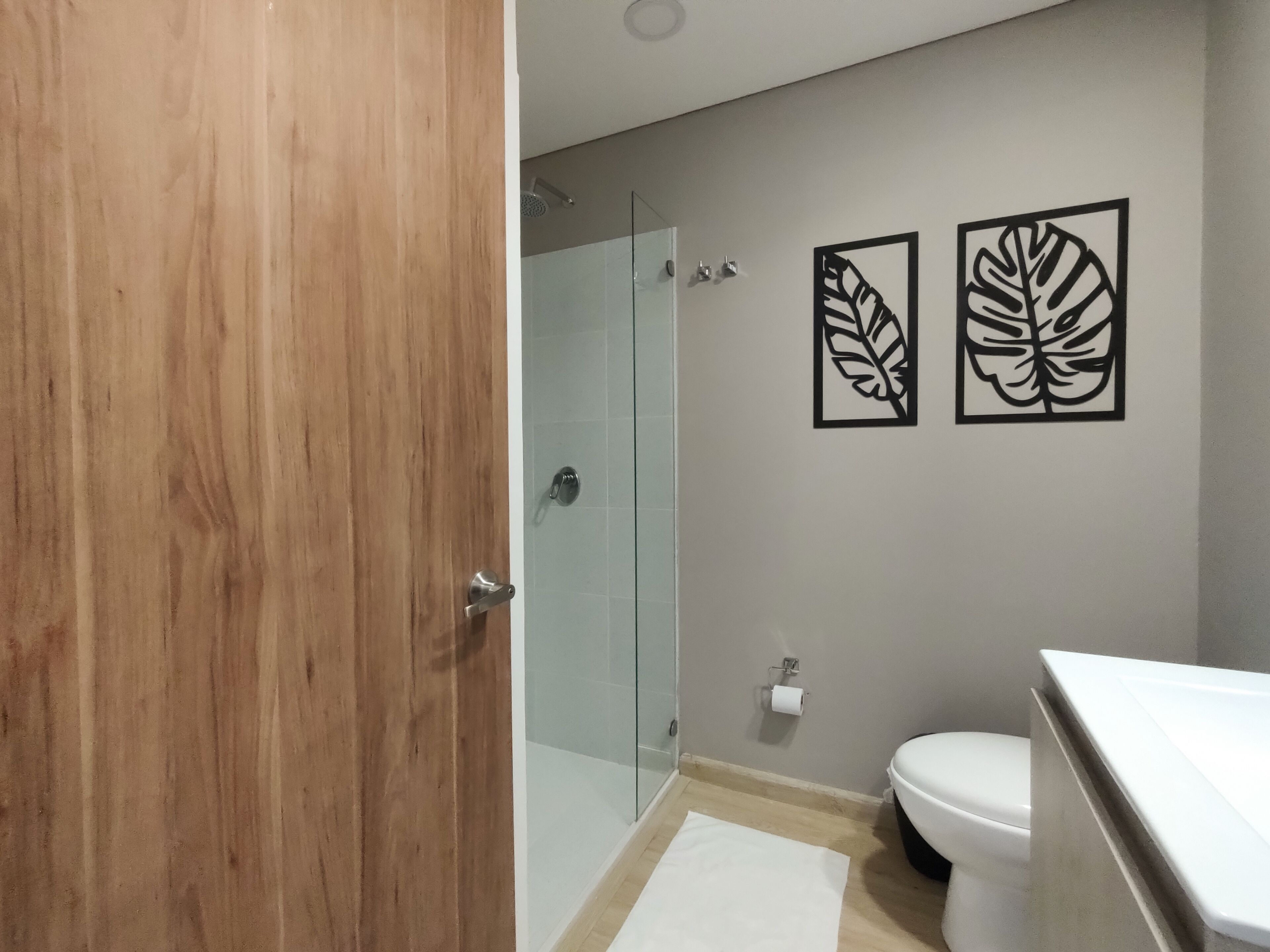 Un baño moderno con puerta de madera, cabina de ducha de vidrio, inodoro y dos obras de arte colgadas en la pared.