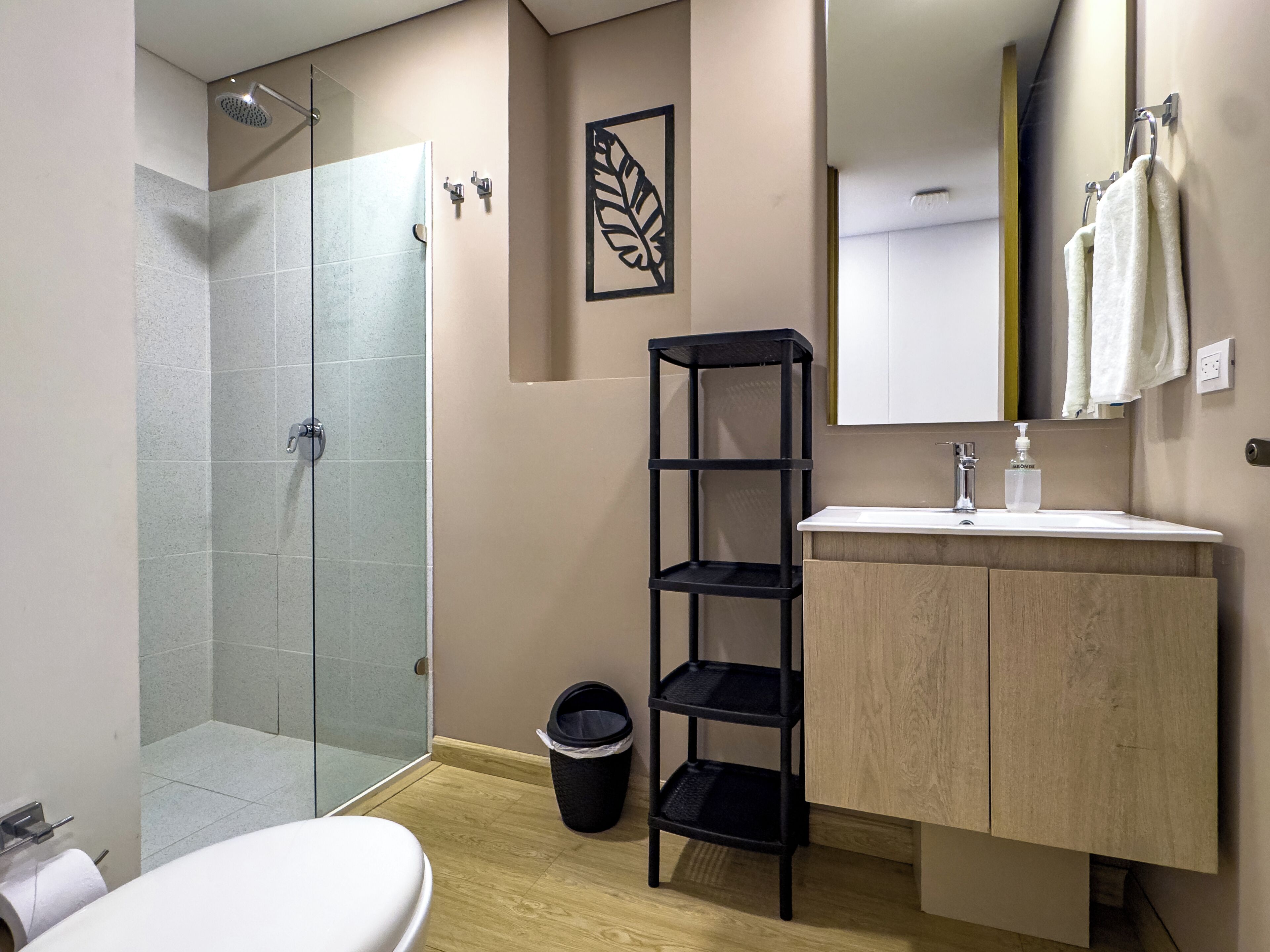 Ein modernes Badezimmer mit einer Duschkabine aus Glas, einem Holzbadezimmermöbel, einem schwarzen Regal und einem Handtuch an der Wand.