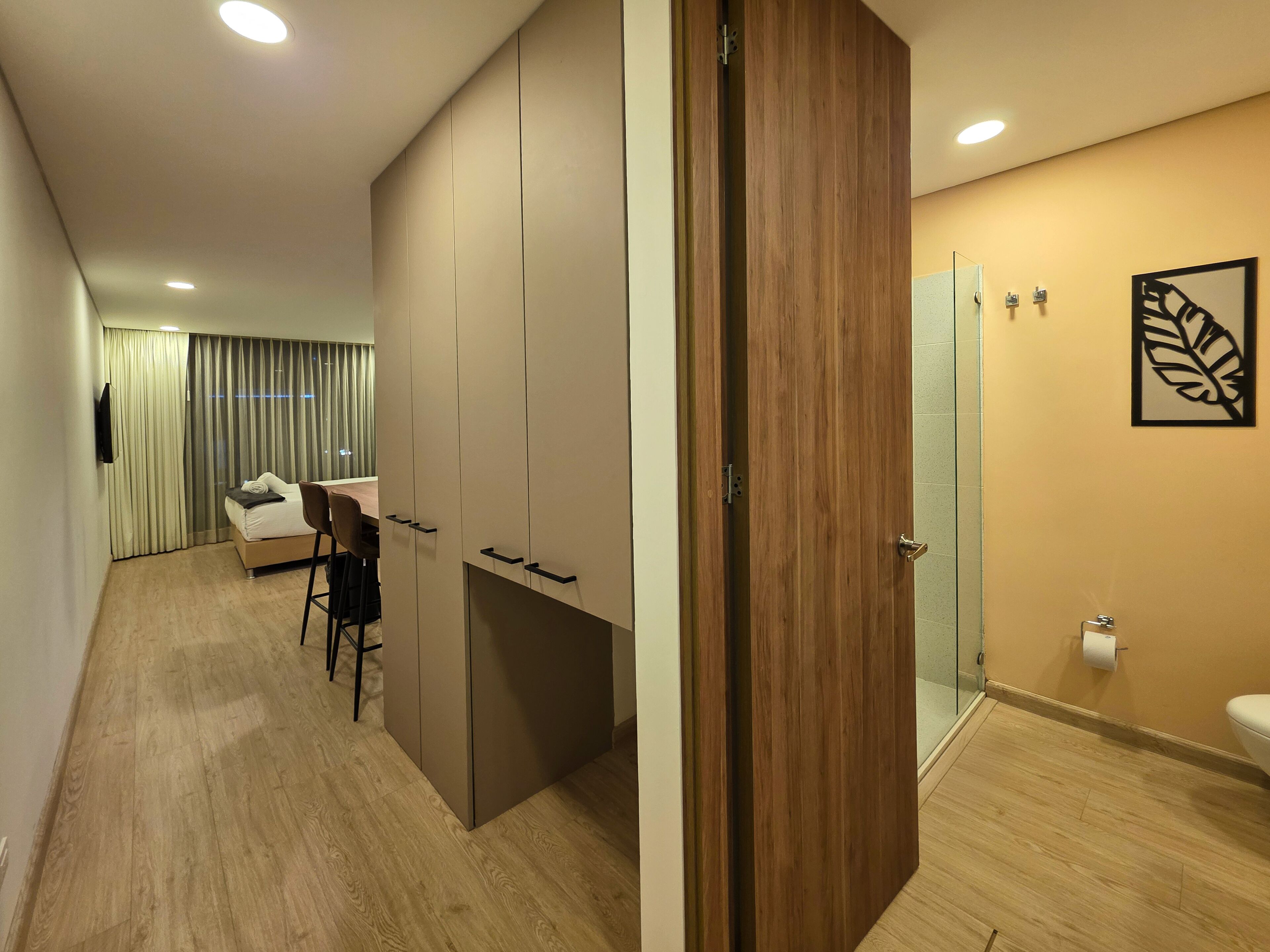 Habitación de hotel moderna con suelo de madera, cama con cabecera gris, escritorio con silla, armario, baño con inodoro y lavamanos, y un gran ventanal con cortinas.