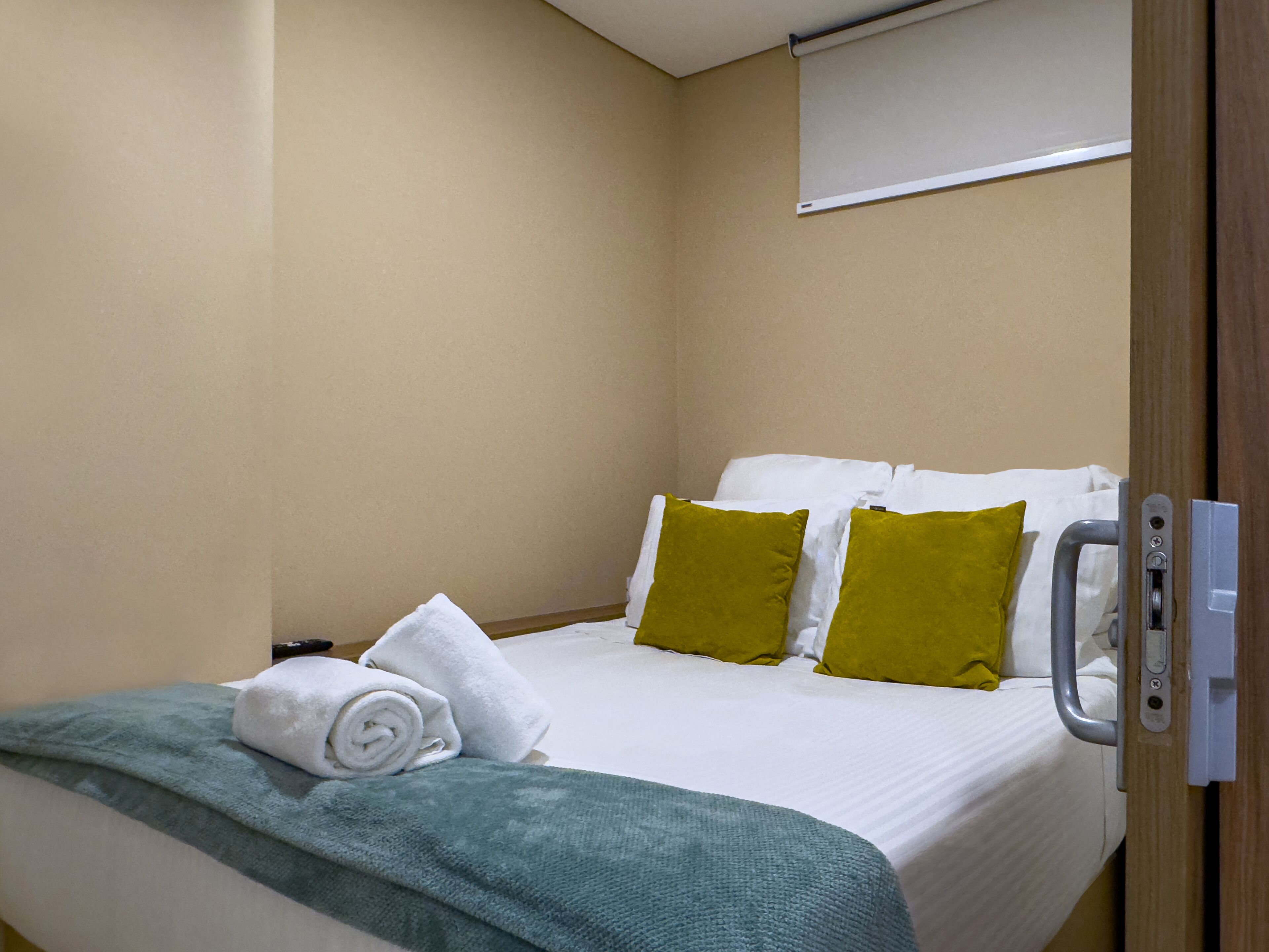 Habitación de hotel con cama, ropa de cama blanca, una manta verde y dos almohadas verdes.