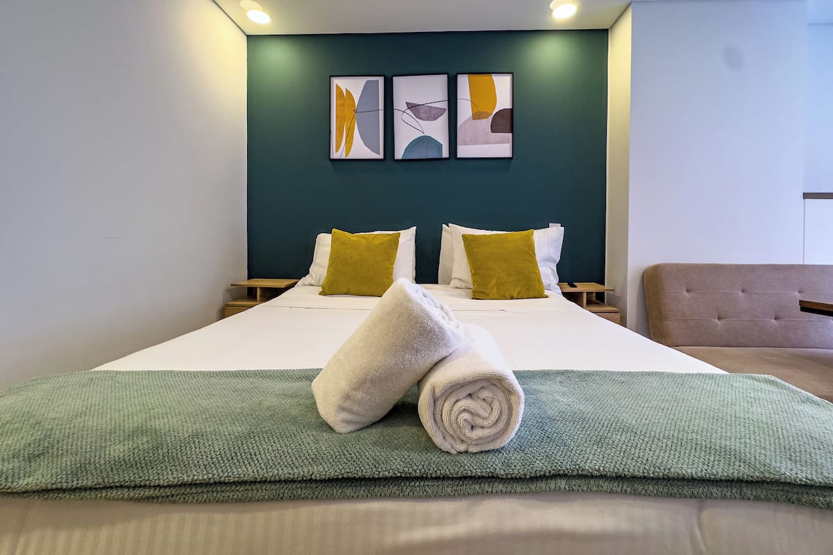 Une chambre d’hôtel avec un lit, deux oreillers, une couverture verte et une serviette enroulée. Il y a deux tables de chevet et une tête de lit.