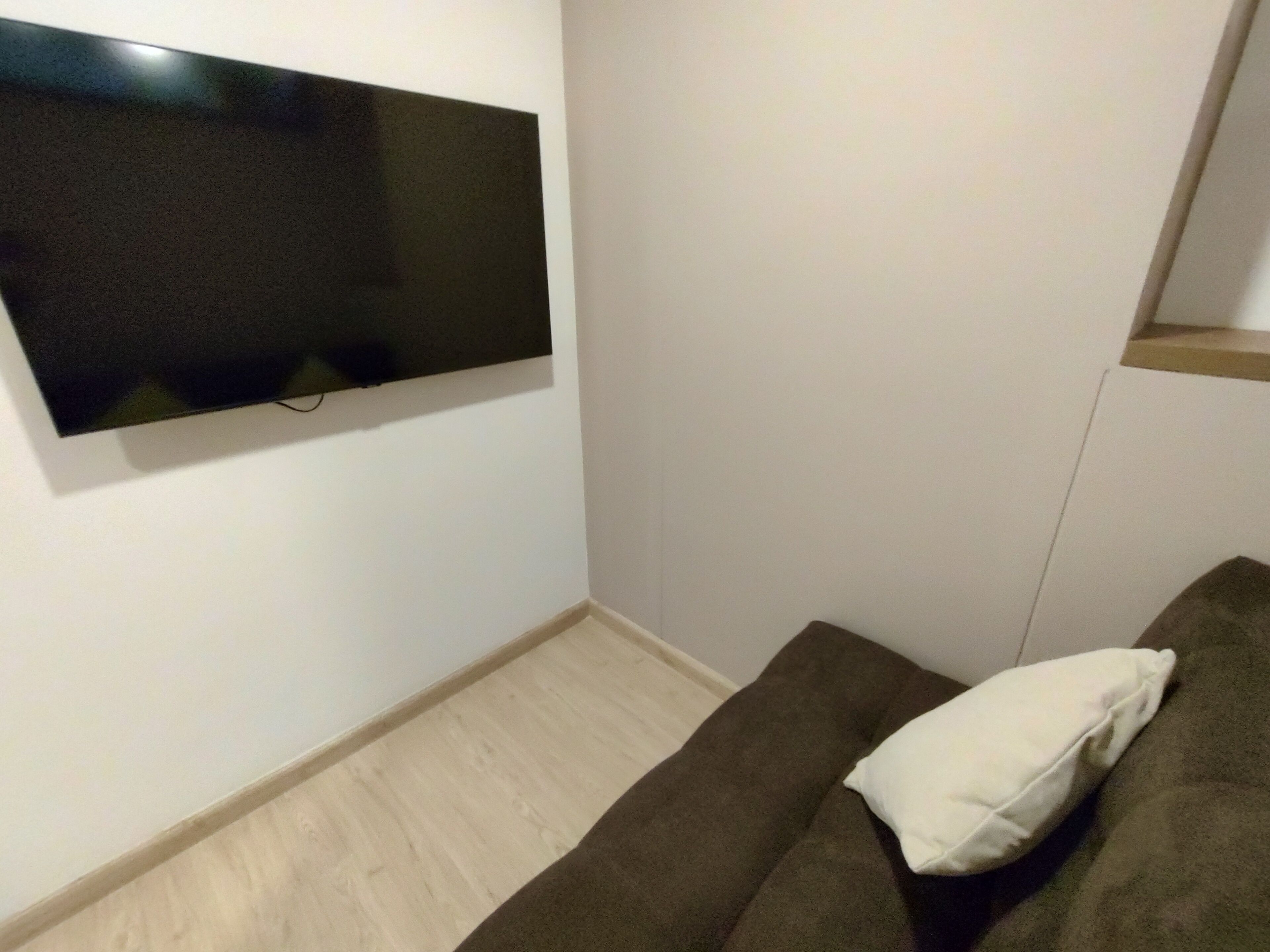 Une pièce dotée d’un téléviseur à écran plat fixé au mur, d’un canapé marron agrémenté d’un coussin blanc et d’un parquet clair.
