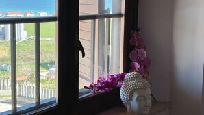Una finestra con vista sul mare e sugli edifici, una statua e fiori viola su un davanzale.
