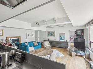 A modern living room with a blue sofa, a round coffee table, and a kitchen area with a large TV. - Quai des Célestins  1bdr by B'Your Home (Paris)