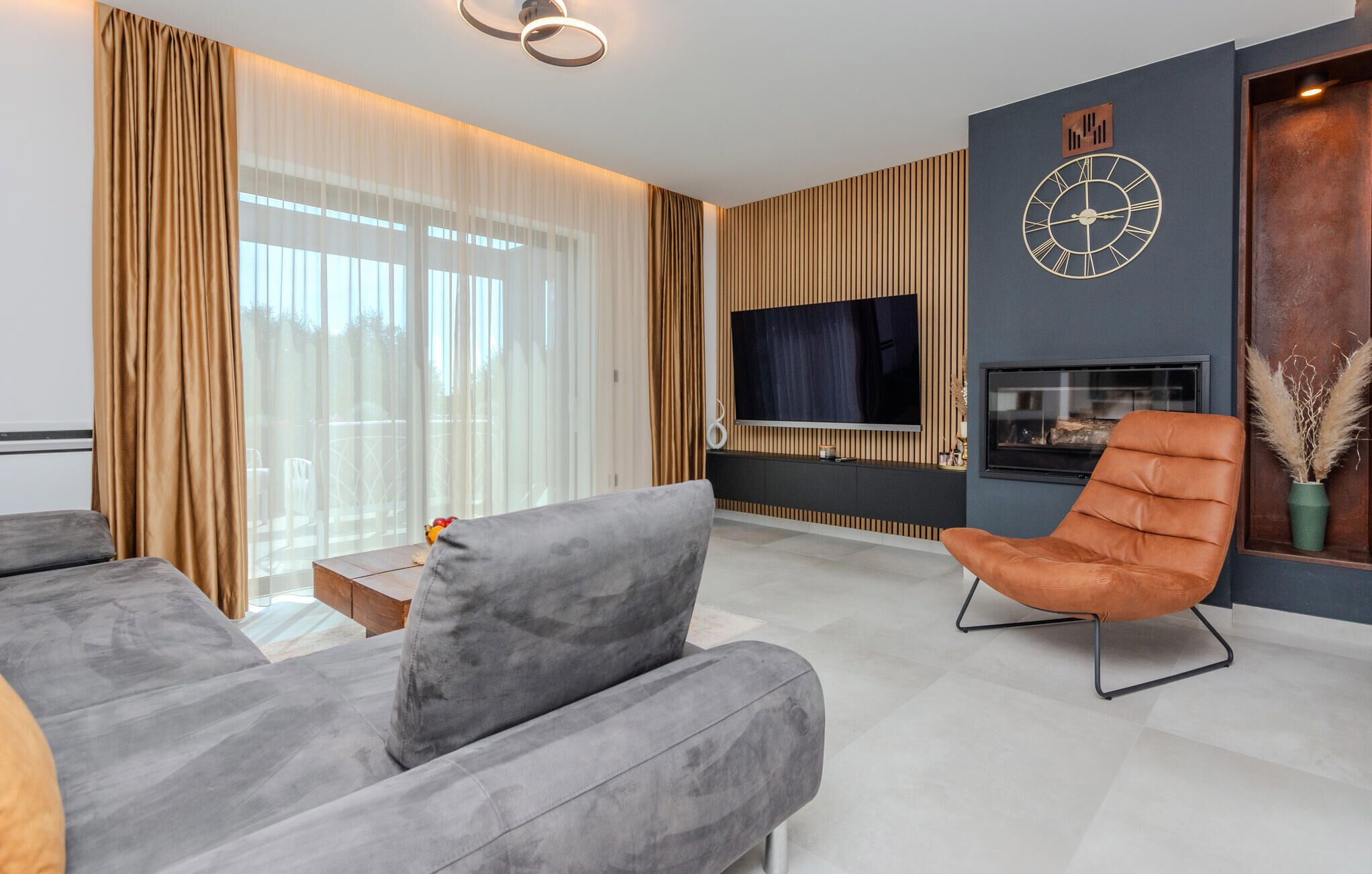A modern living room with a grey sofa, a brown leather armchair, a flat-screen TV mounted on a wooden wall, and a fireplace.