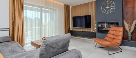 A modern living room with a grey sofa, a brown leather armchair, a flat-screen TV mounted on a wooden wall, and a fireplace.