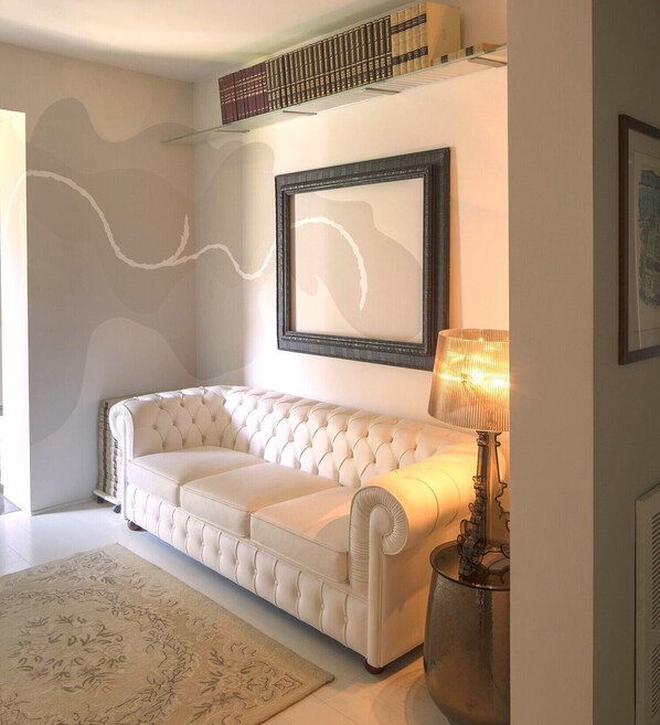 A white tufted sofa in a well-lit room with a patterned rug, a framed abstract painting, and a lamp on a side table.