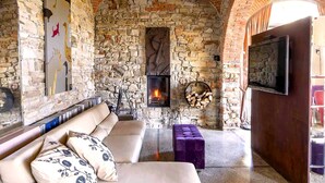 A cozy room with a stone wall fireplace, a sofa with patterned cushions, and a television mounted on the wall. - Tenuta Moncarlino - room Terra (Piemonte)