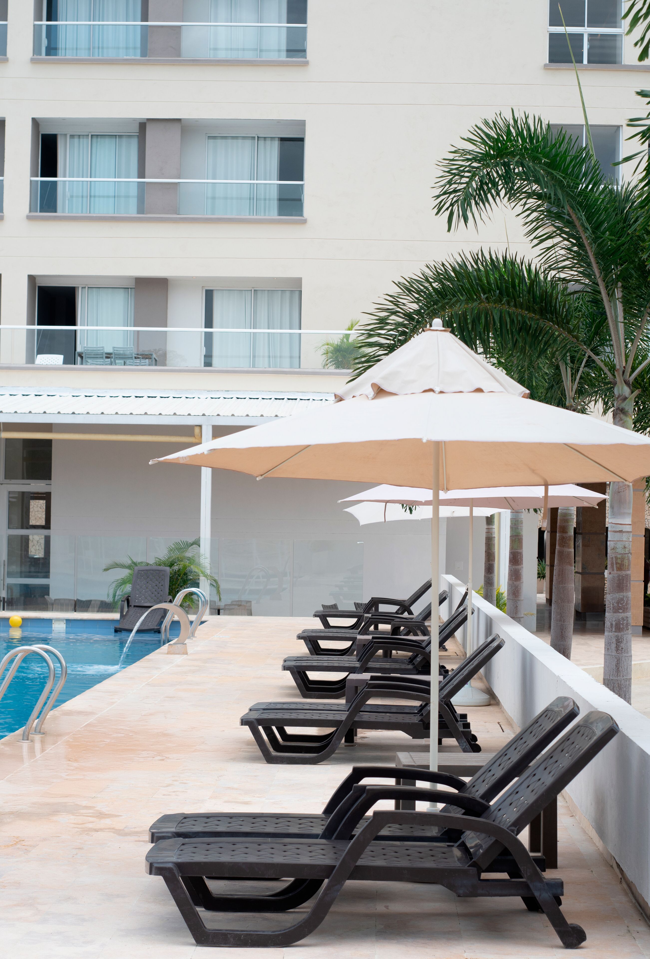A poolside area with lounge chairs and umbrellas, a pool, and a building with balconies in the background.