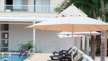 A poolside area with lounge chairs and umbrellas, a pool, and a building with balconies in the background.