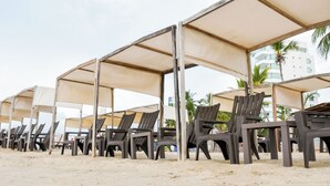 Rows of beach chairs under canopies, with a sandy beach and palm trees in the background.