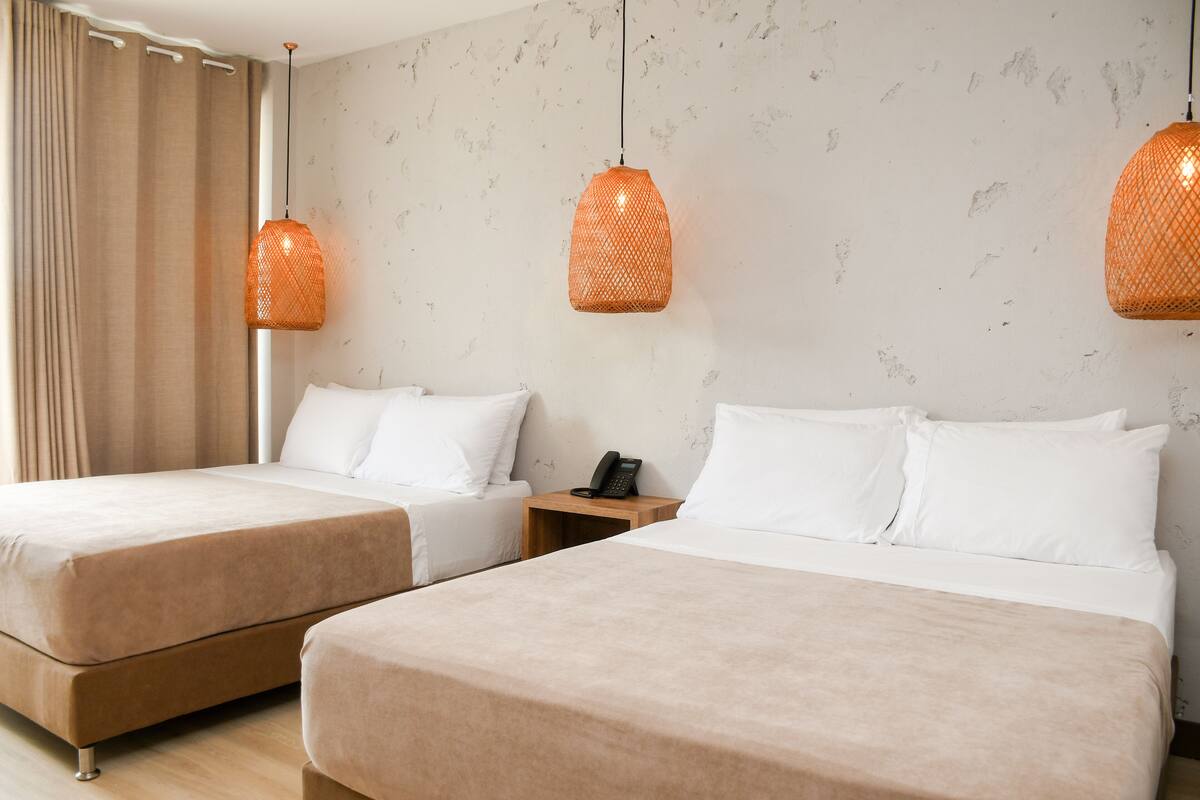 A hotel room with two beds, beige bedding, a wooden nightstand with a phone, and orange pendant lights.