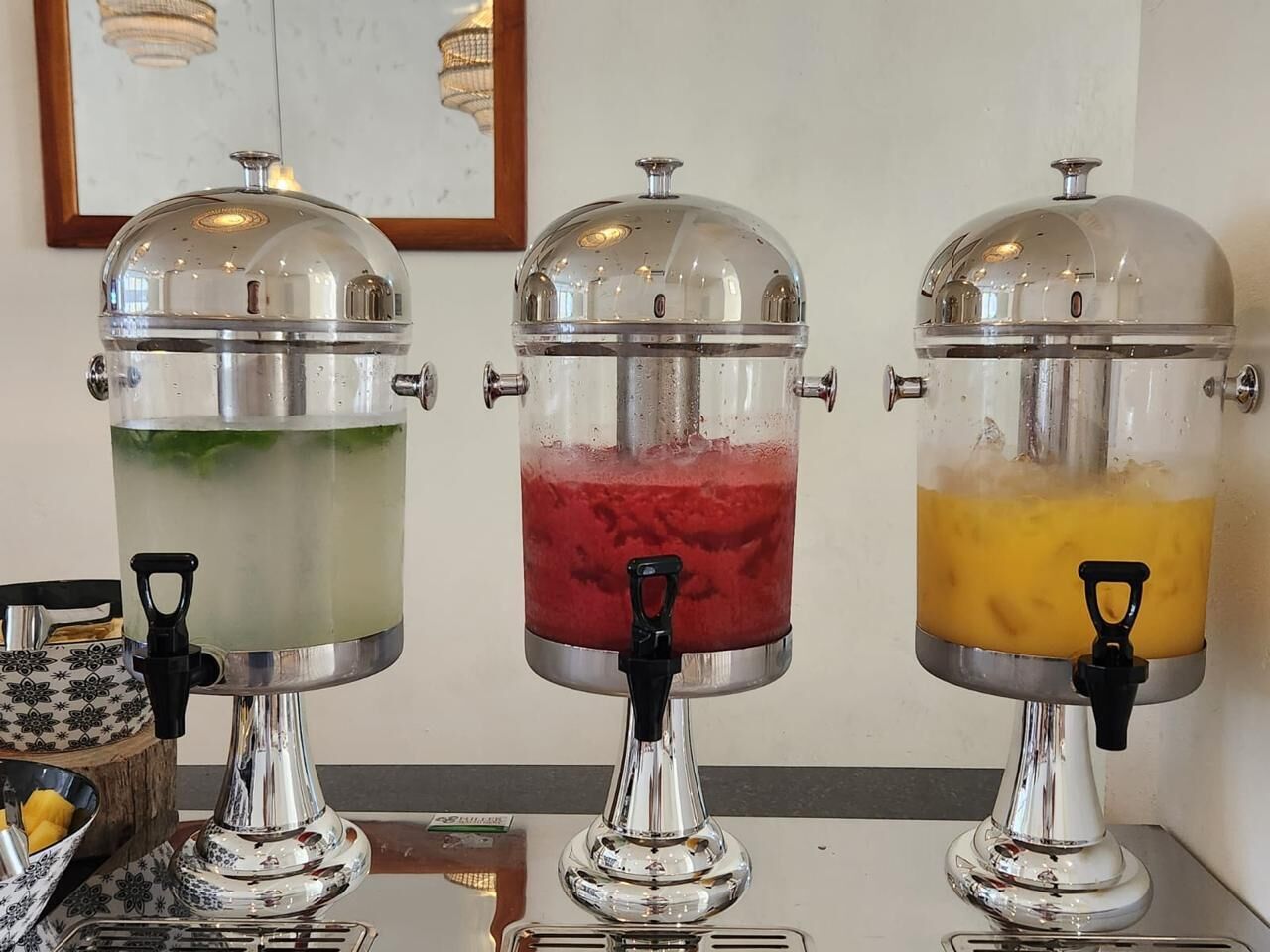 Tres dispensadores de bebidas con contenedores transparentes y bases metálicas, cada uno con un asa negra.