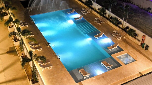 An aerial view of a rectangular swimming pool with multiple lounge chairs and a small hot tub area, surrounded by palm trees and a paved walkway.