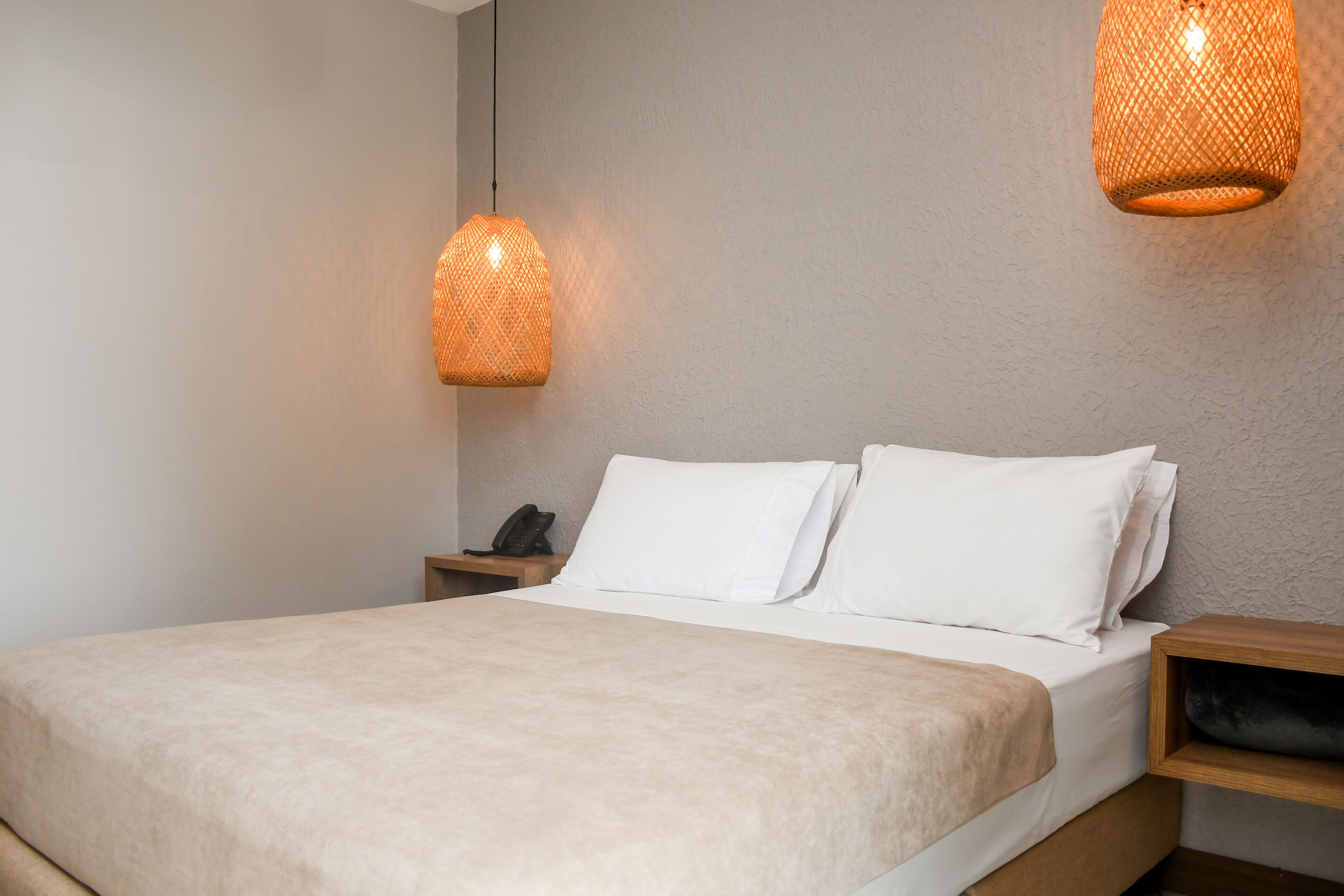 A neatly made bed with white linens and a beige throw blanket, a bedside table with a phone, and two hanging lamps with a woven design.