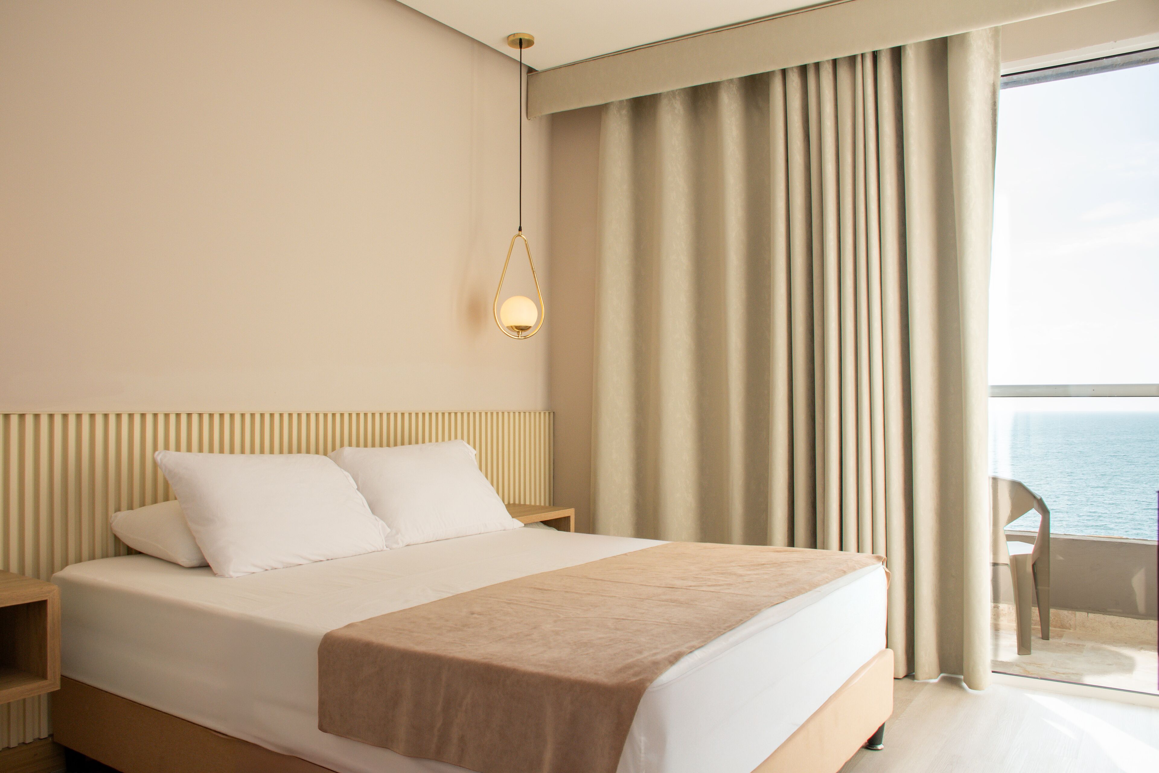 A modern hotel room with a large bed, a bedside table, a hanging light fixture, and a view of the sea through the window.